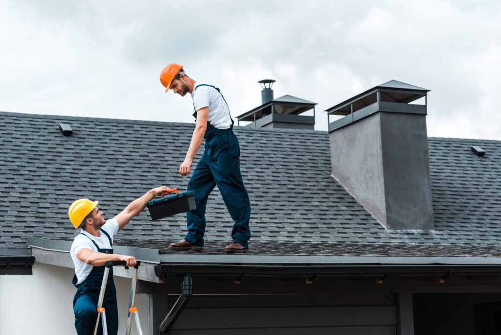 Roofing Services Lynn Haven, FL handyman in helmet giving toolbox to handsome colleague standing on rooftop