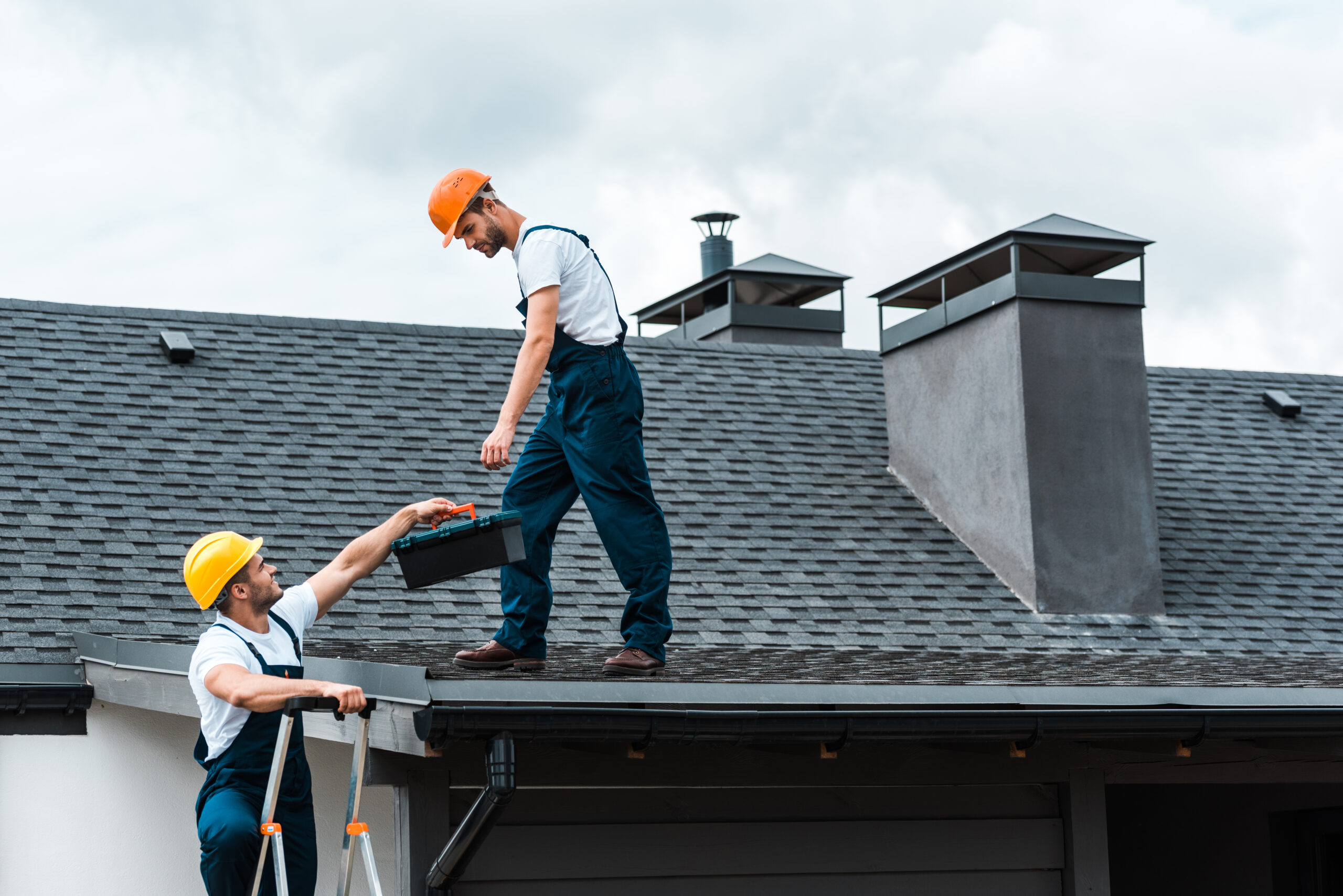 Roofing Services Lynn Haven, FL handyman in helmet giving toolbox to handsome colleague standing on rooftop