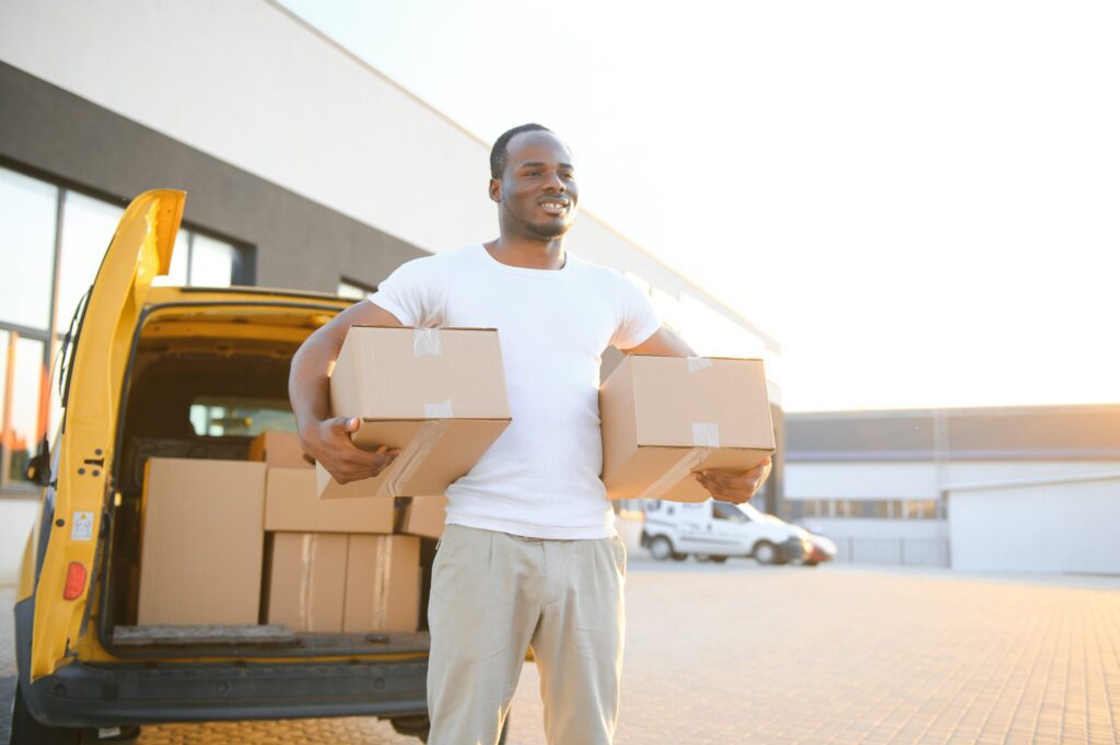 Moving Company in Yonkers, NY Portrait of a young African American package delivery man near a car with boxes.